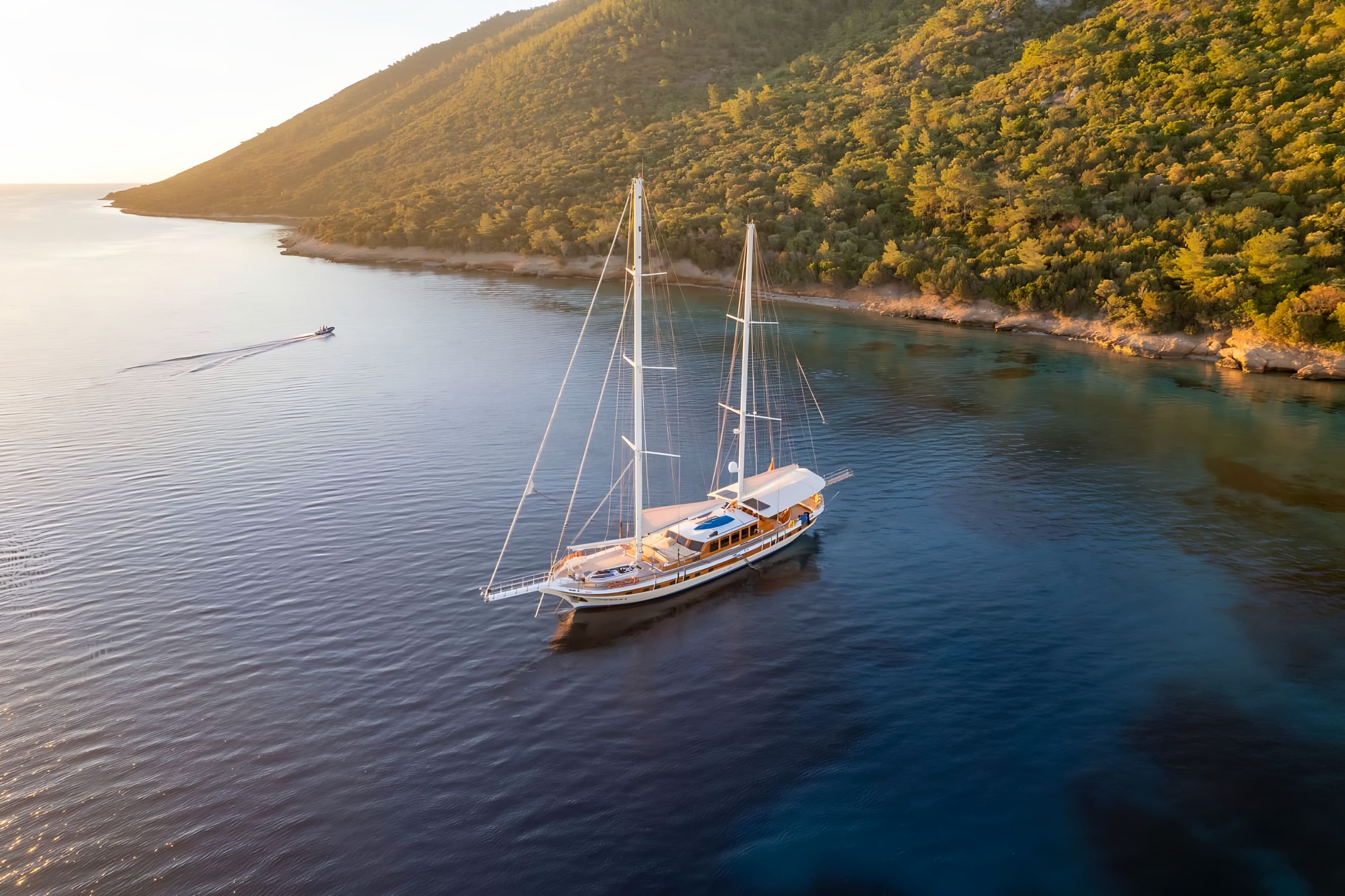 A two-masted wooden gulet at anchor in a calm bay at sunset, below a pine-covered hillside